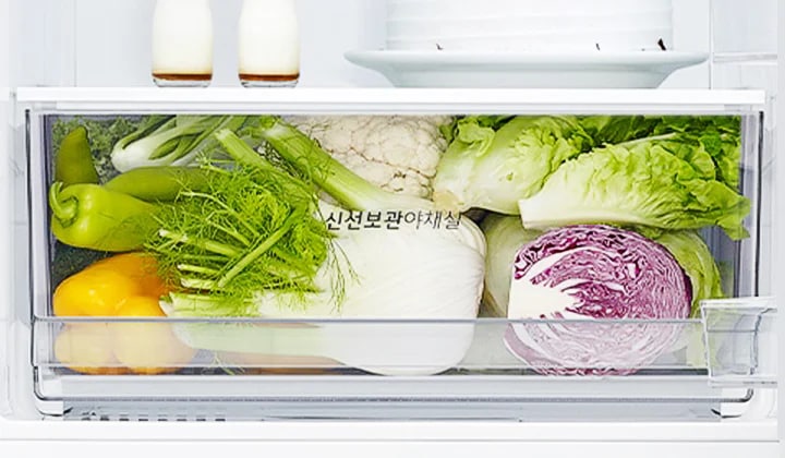 냉장고 내부의 채소 보관 칸에 다양한 채소들이 가득 담겨 있습니다. 노란색과 초록색 피망, 양배추, 브로콜리, 상추, 적양배추 등이 보이며, 채소 칸에는 신선보관야채실이라는 문구가 적혀 있습니다. 위쪽 선반에는 작은 병 두 개와 접시가 놓여 있습니다.